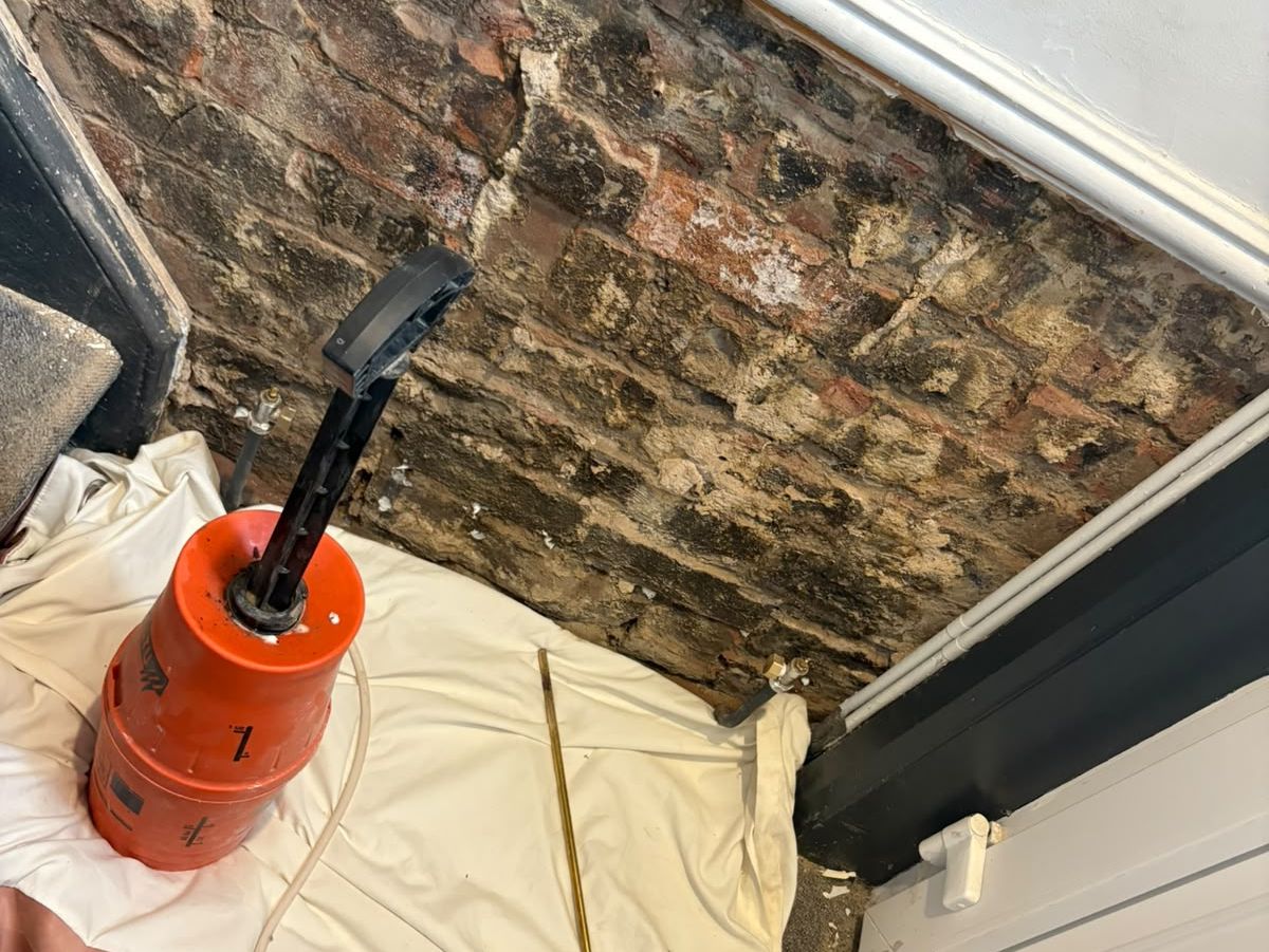 A pressurised chemical damp proof course injection vessel and lance beside exposed, salt-stained brickwork
